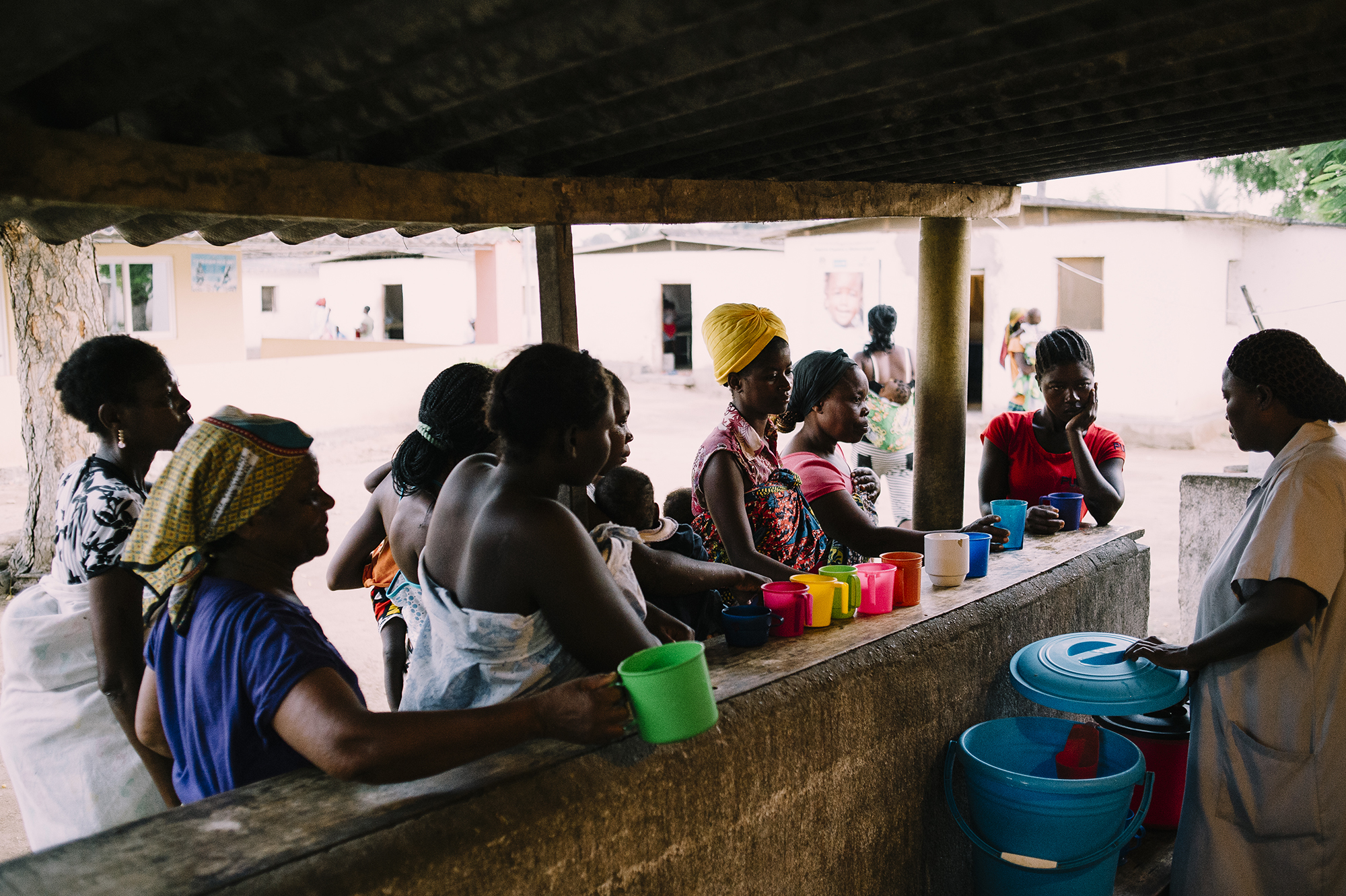 Mothers in a malnutrition clinic in Angola receiving therapeutic milk, engaging in community support, with colorful cups lined up on a counter.