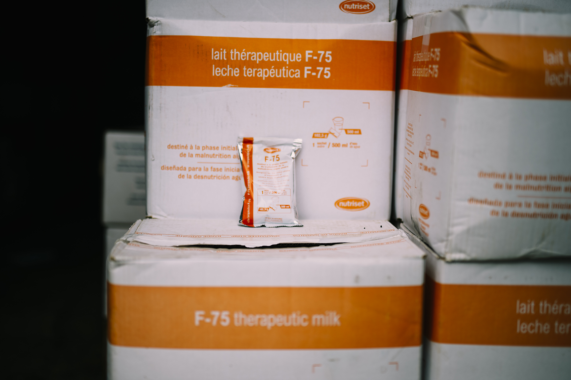 Therapeutic milk packets labeled F-75 on stacked boxes in a malnutrition clinic, highlighting nutritional support for infants in Angola.