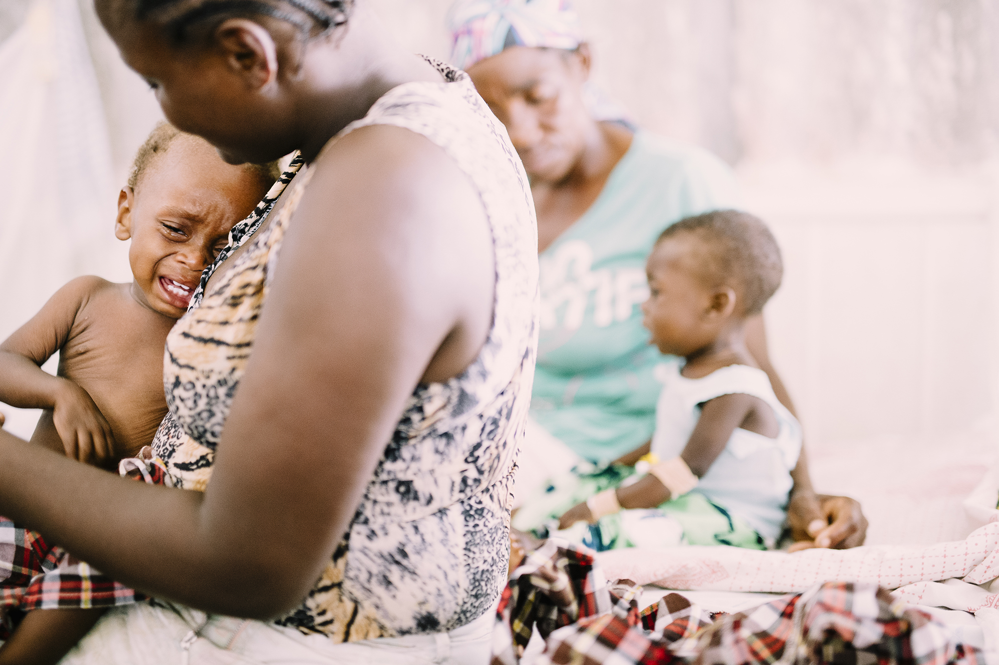 Mother comforting malnourished baby in clinic, surrounded by other mothers and children, emphasizing emotional support and care in malnutrition treatment.