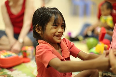 Smiling young girl in a red shirt engaged in play with toys, surrounded by children in a supportive environment, illustrating the joy and care provided by Saving Moses' NightCare program.