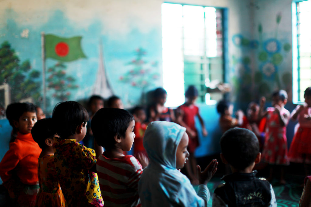 Children in colorful clothing gathered in a classroom with a mural of the Bangladeshi flag, highlighting the need for safe spaces for vulnerable children in Bangladesh.