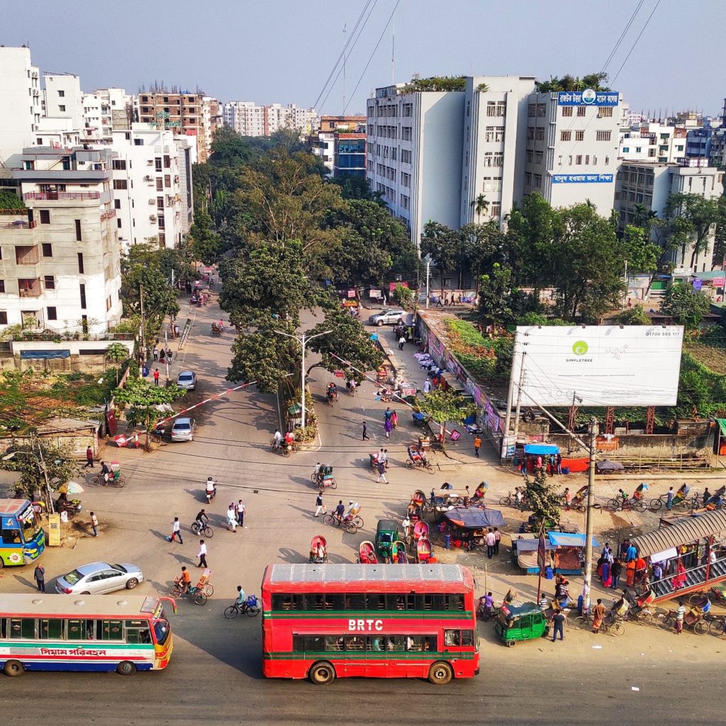 Busy street scene in Dhaka, Bangladesh, featuring a red BRTC bus, various vehicles, pedestrians, and vendors amidst dense urban buildings and greenery, illustrating the city's overcrowded environment.