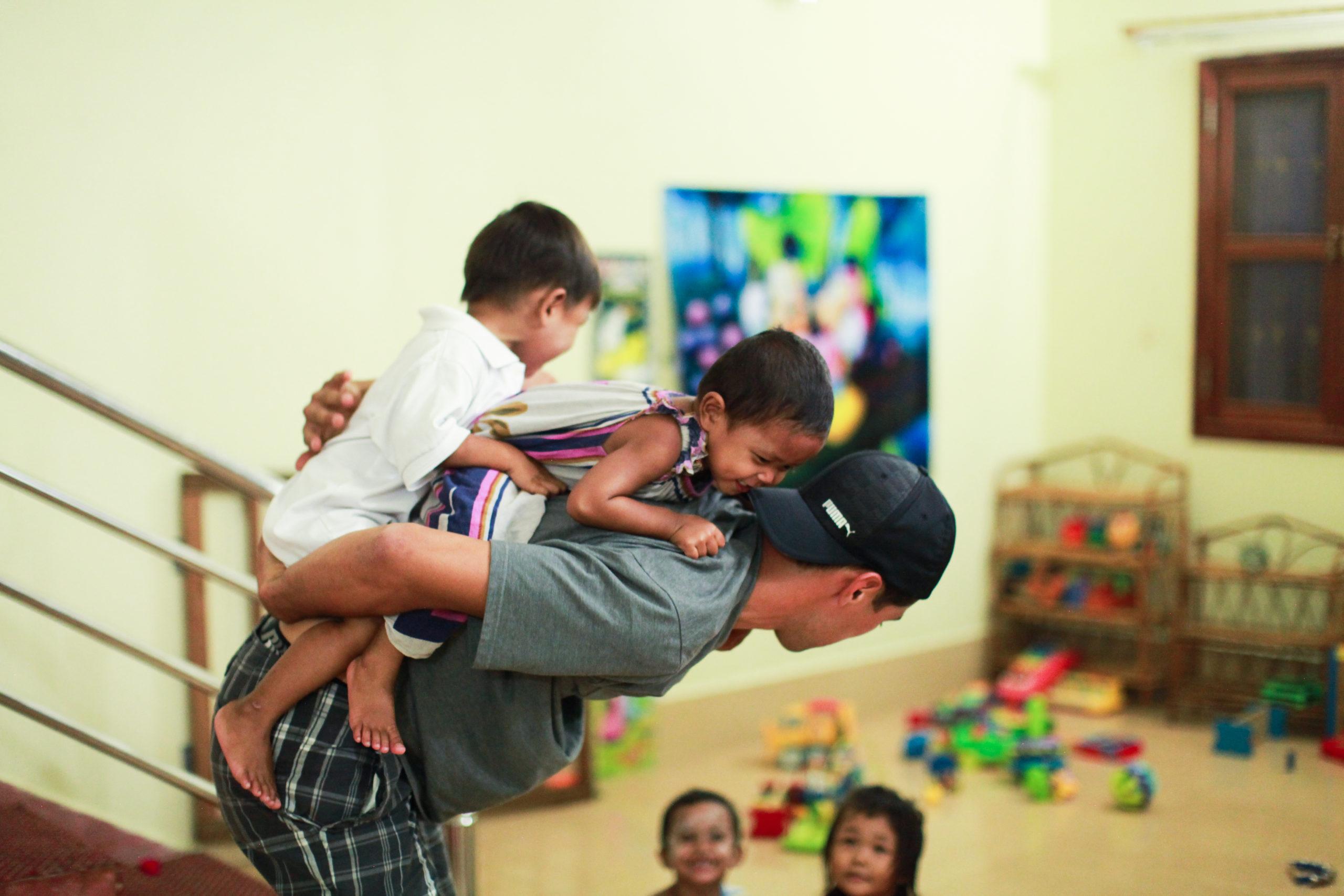 Child playing on adult's back in a colorful playroom, with toys scattered on the floor and a vibrant mural in the background.