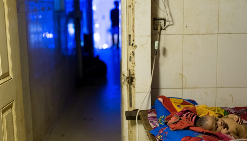 Baby sleeping on a colorful blanket in a dimly lit room, with a blurred figure in the background, representing the vulnerable children cared for by the NightCare program for sex workers.