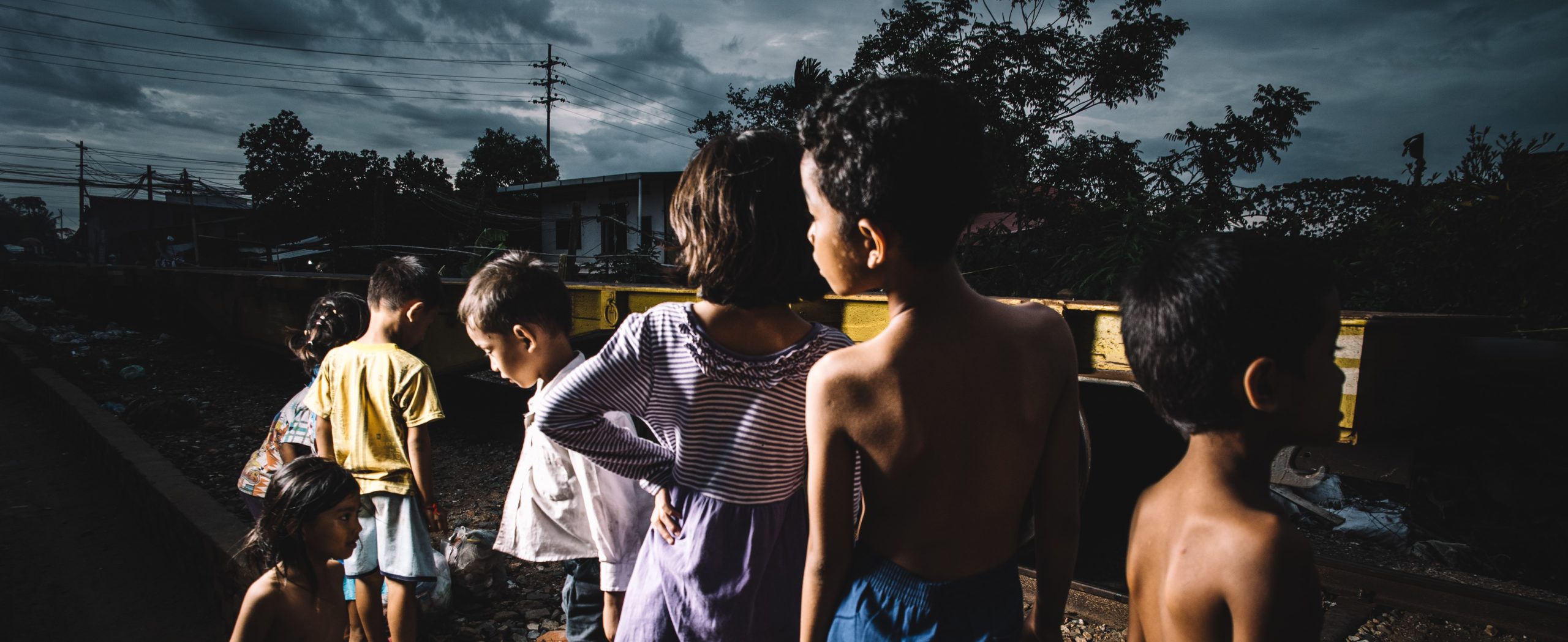 Group of children in impoverished setting, highlighting issues of child human rights, facing away from the camera, with a backdrop of urban decay and cloudy skies.