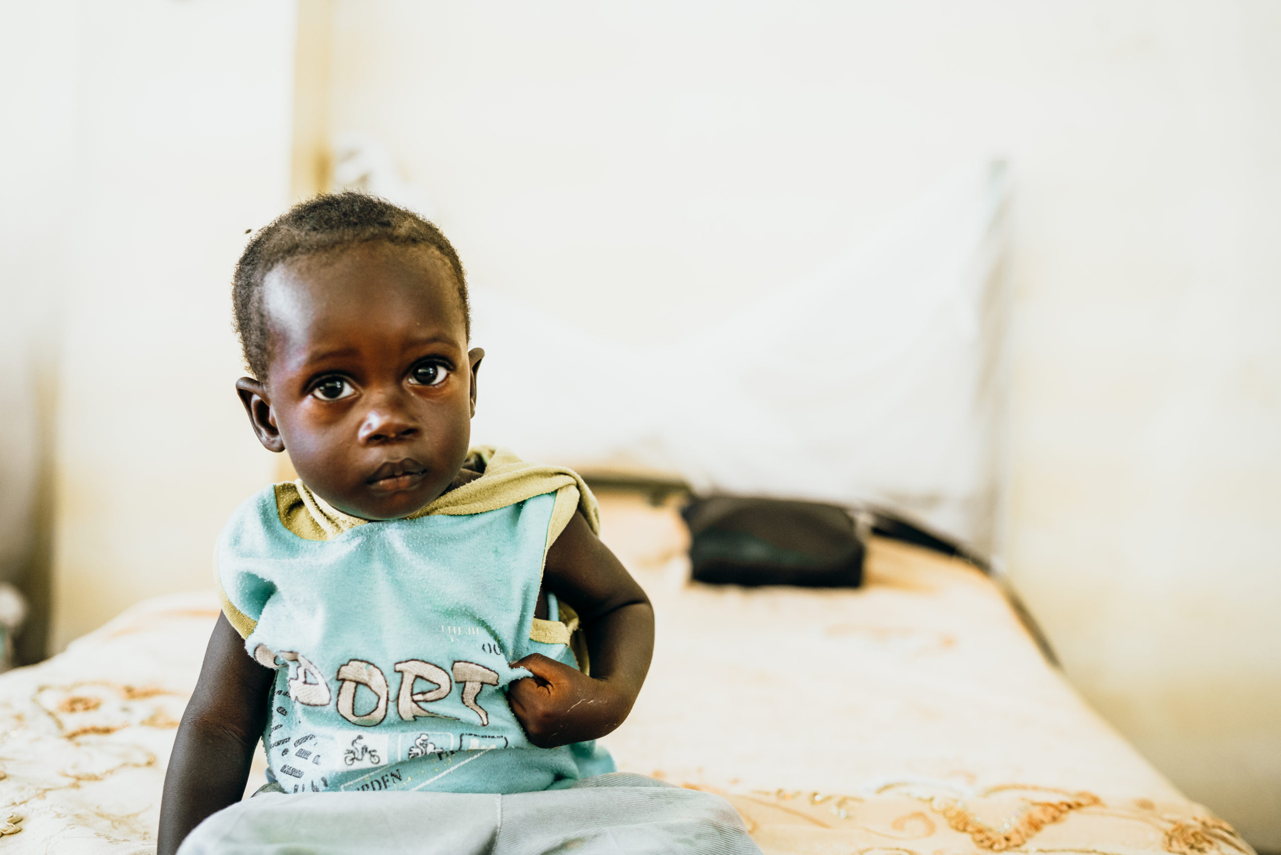 Child sitting on a bed, showing signs of malnutrition, highlighting the urgent need for nourishment and support for infants in developing countries.