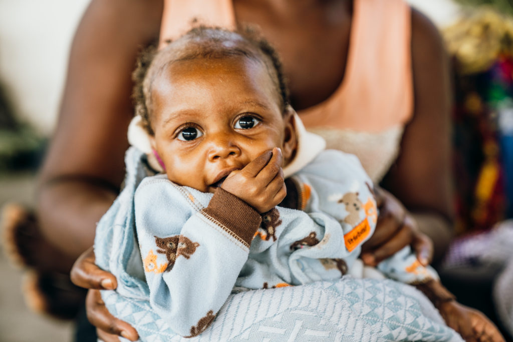 Baby with big, expressive eyes sucking on a finger, wearing a cozy blue outfit, held by a caregiver, symbolizing the vulnerable infants supported by Saving Moses' humanitarian efforts.