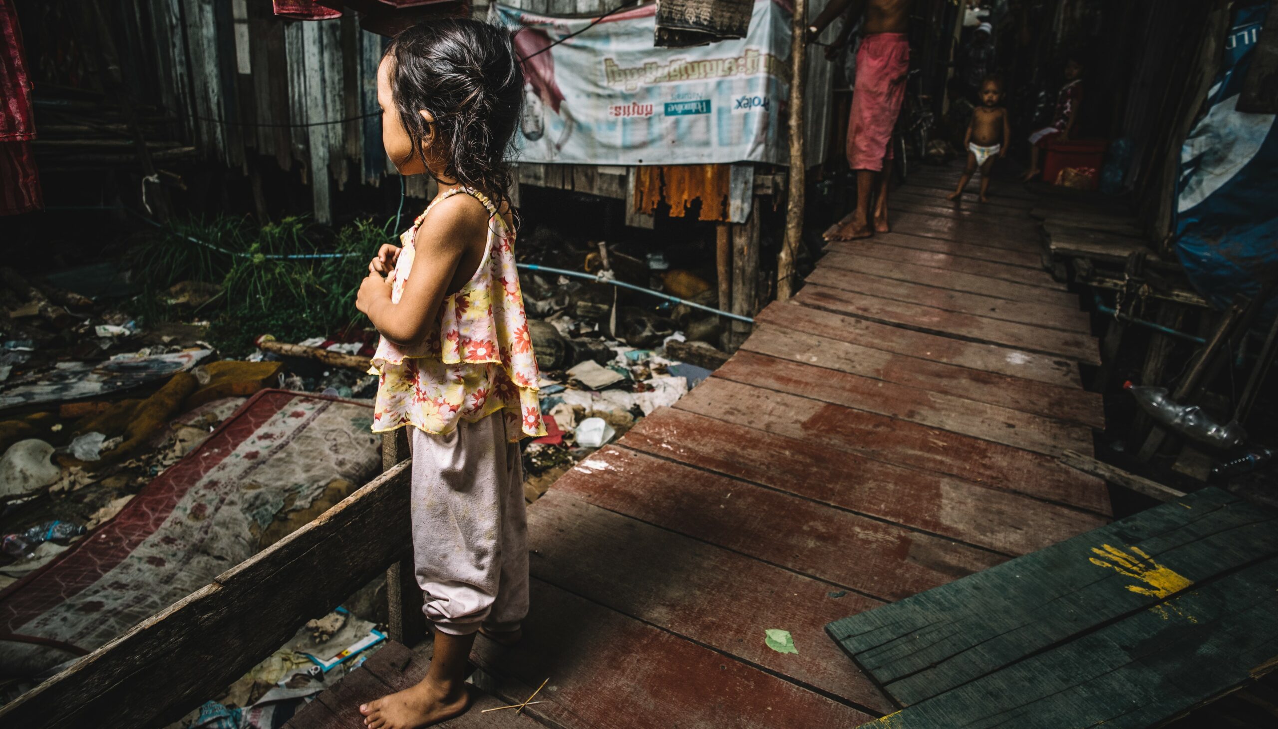 Young girl standing on a wooden walkway in a slum area, surrounded by litter and debris, highlighting the urgent need for COVID-19 relief efforts for vulnerable children.