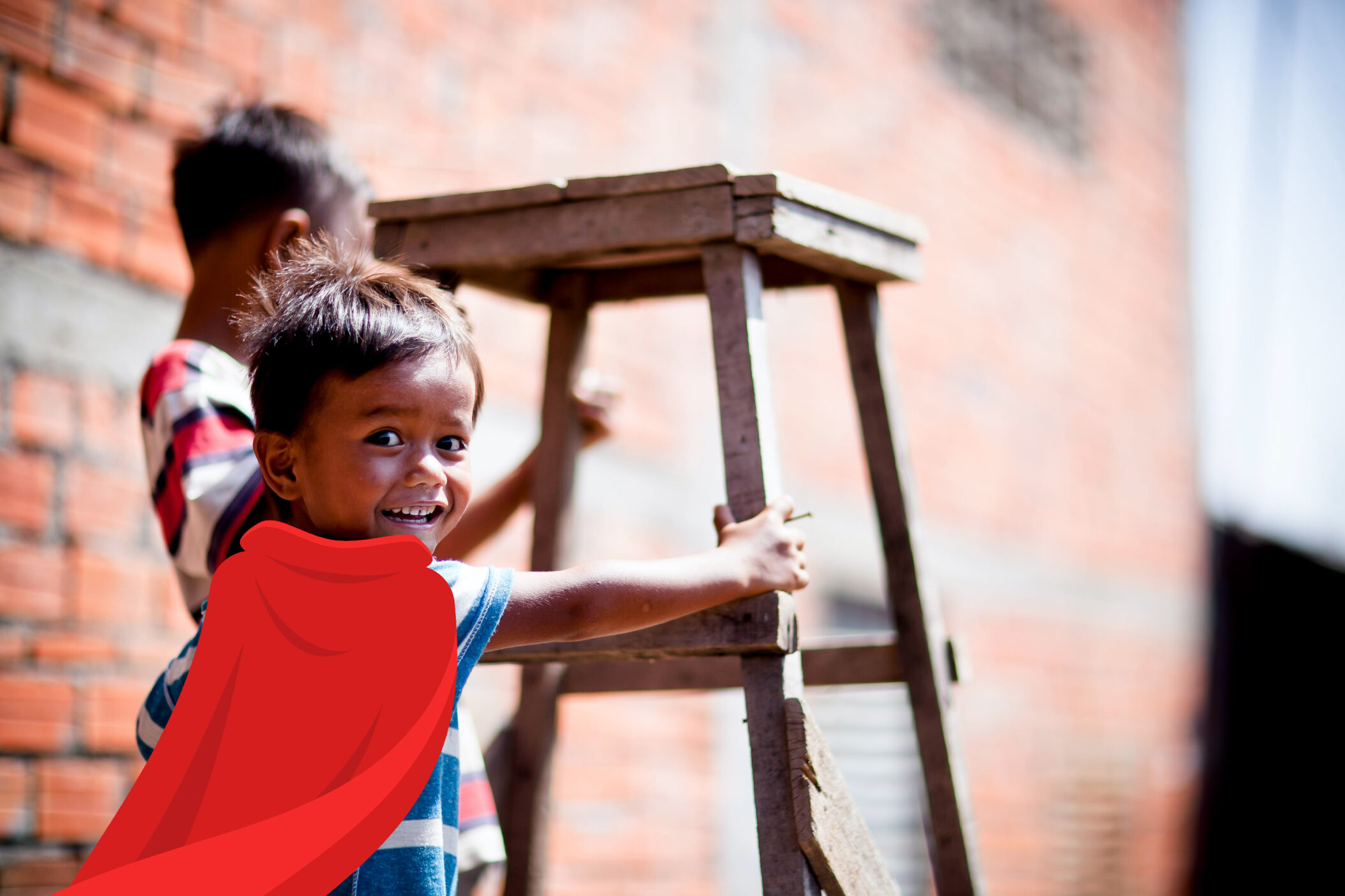 Smiling child in a striped shirt wearing a red cape, standing next to a wooden stool, symbolizing heroism and support for vulnerable children through donations.