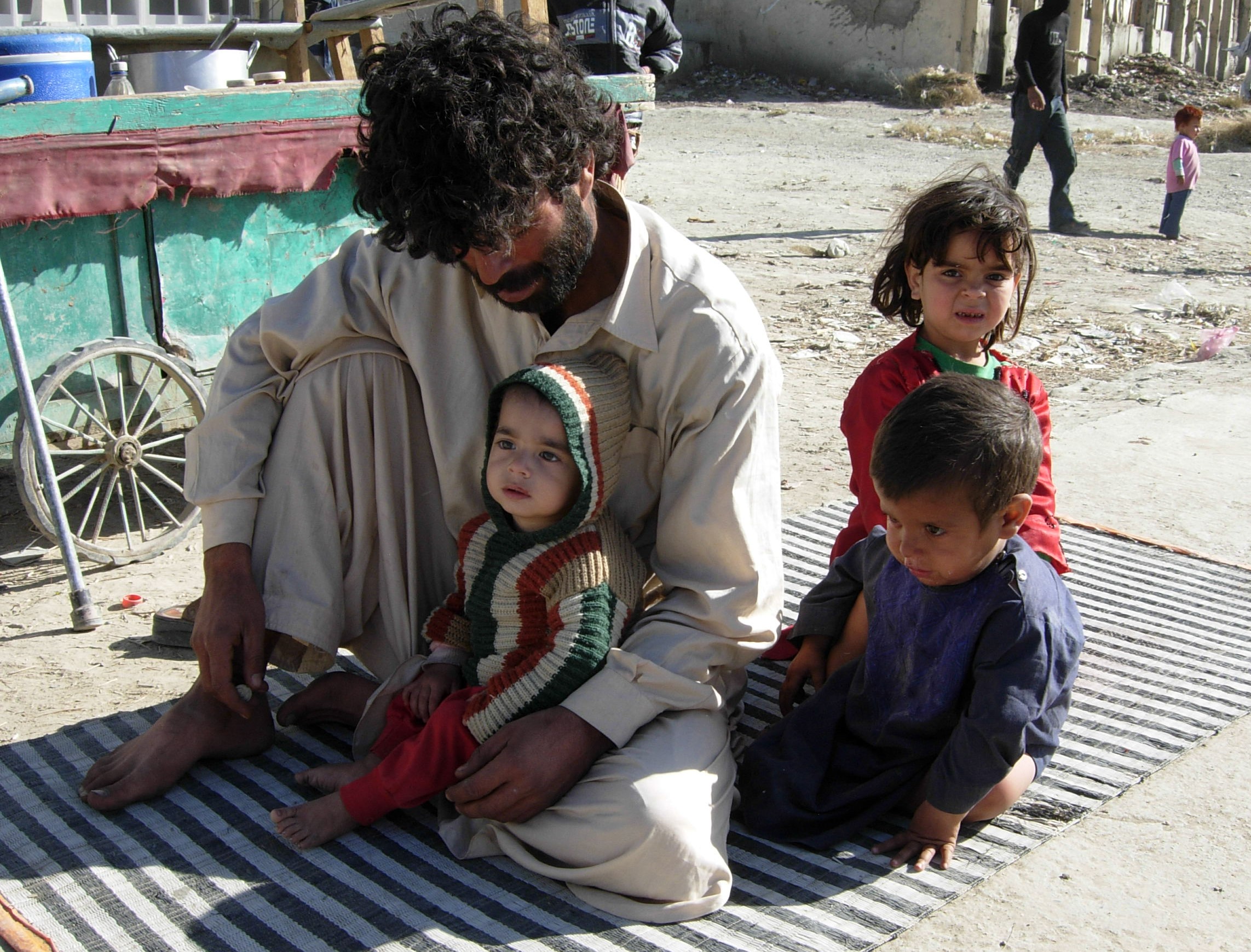 Man sitting on a rug with two children in Afghanistan, one child wearing a striped sweater and the other in a blue outfit, highlighting the challenges faced by families amid the humanitarian crisis.