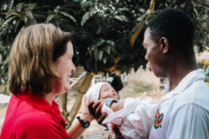 Woman in red shirt smiling at man holding a newborn baby, outdoors with greenery in the background, highlighting the mission of Saving Moses to support vulnerable infants.