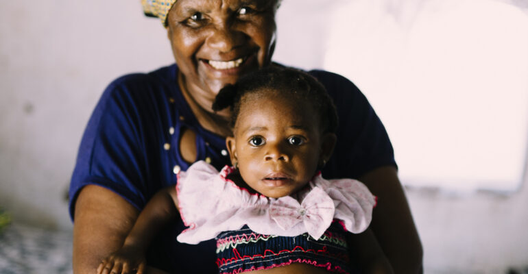 Smiling woman holding a baby, both dressed in colorful clothing, representing the mission of Saving Moses to provide care and support for vulnerable children.