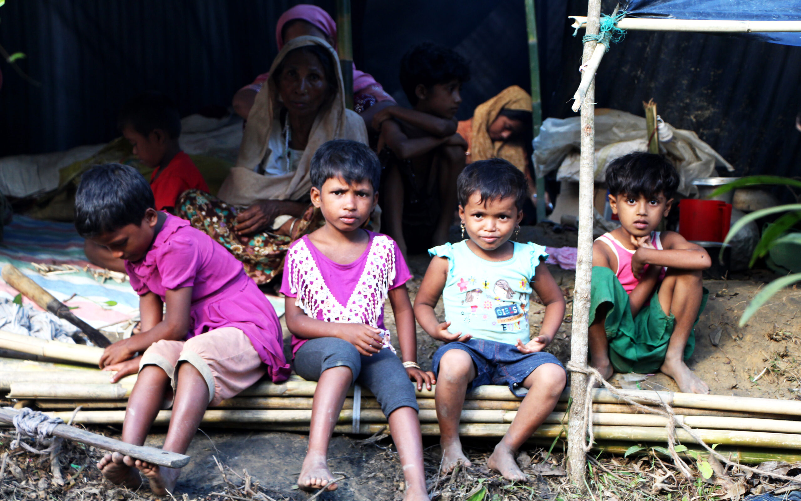 Children sitting on bamboo in a makeshift shelter, reflecting vulnerability and resilience in a humanitarian context, related to discussions on mass violence and dehumanization by Saving Moses.