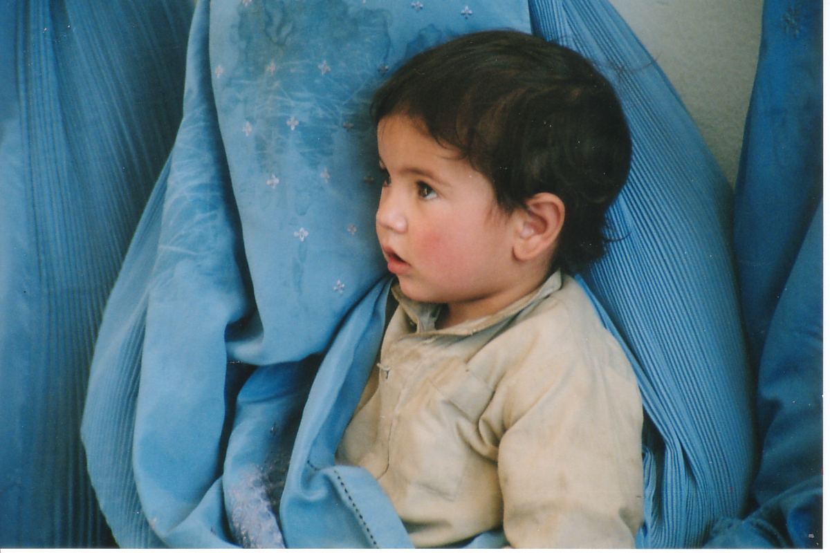 Young Afghan child in traditional clothing, surrounded by blue burqas, reflecting the impact of Taliban rule on women and children in Afghanistan.