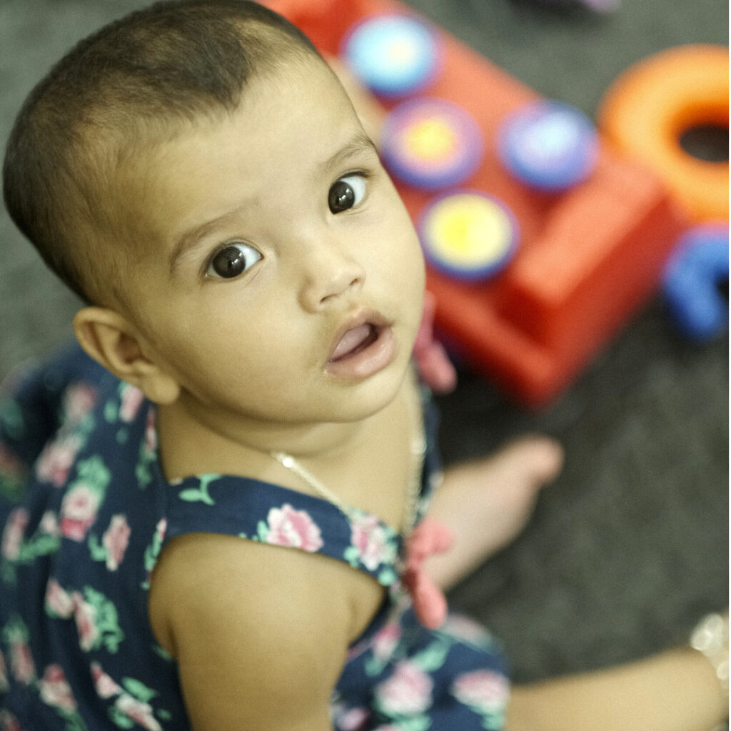 Baby girl with short hair and floral dress, looking up with curiosity, surrounded by colorful toys, representing vulnerable children supported by Saving Moses.
