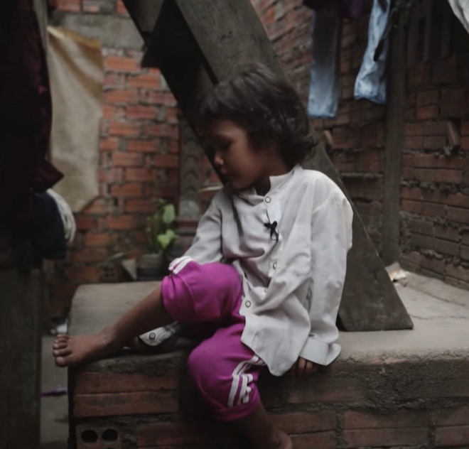 Child sitting on a ledge in a poverty-stricken environment, wearing a white shirt and pink pants, reflecting the impact of malnutrition and hardship.