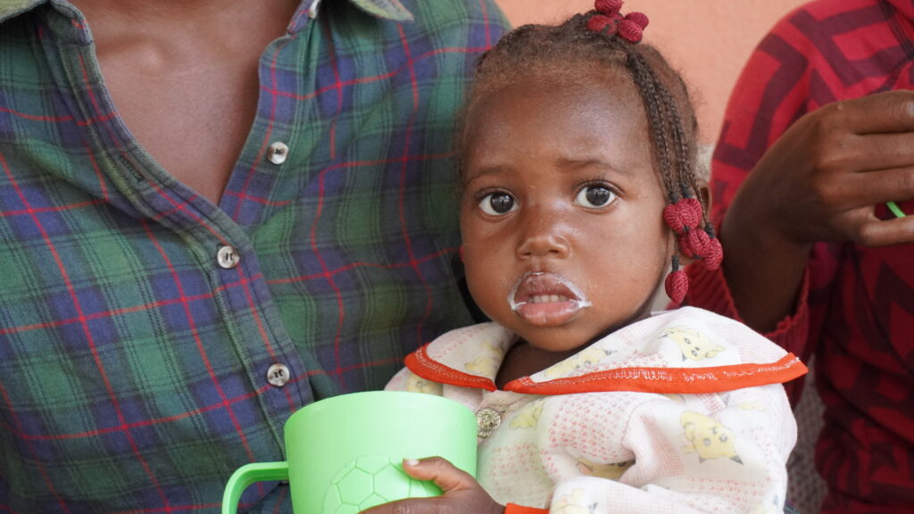 Child with malnutrition symptoms, holding a green cup, displaying milk around mouth, seated next to caregiver, highlighting the impact of Severe Acute Malnutrition.