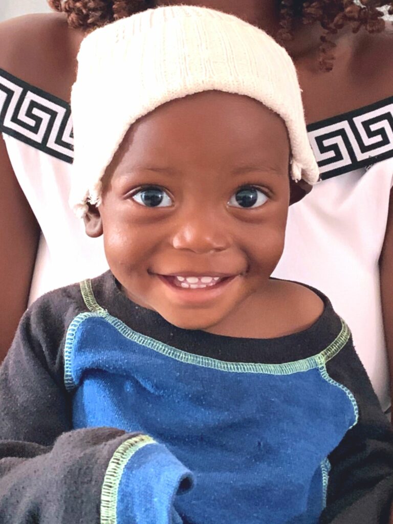 Smiling toddler wearing a white knit hat and blue shirt, representing the impact of Saving Moses' mission to support vulnerable children globally.