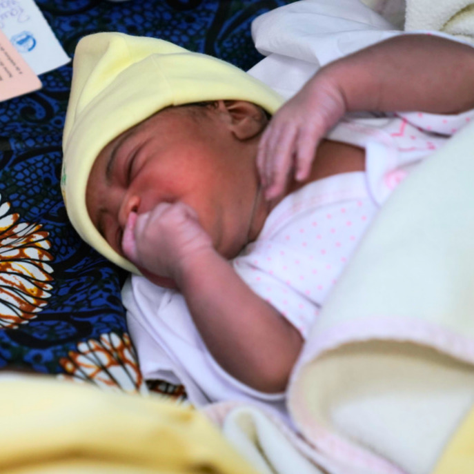 Newborn baby in yellow hat, lying on patterned fabric, highlighting vulnerability and care in conflict zones, relevant to infant mortality and healthcare challenges.