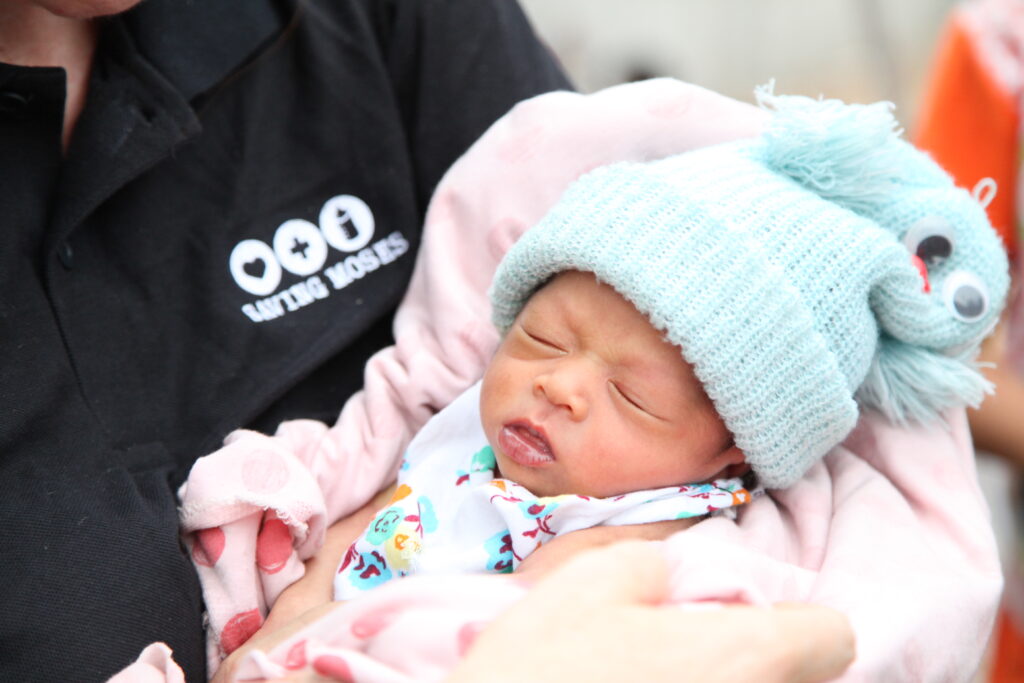 Baby wrapped in a pink blanket, wearing a blue knitted hat with eyes, held by a caregiver in a Saving Moses shirt, symbolizing care for vulnerable infants in conflict zones.