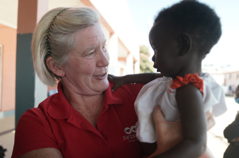 Woman in red shirt from Saving Moses holding and smiling at a young child in a white dress, emphasizing care and connection in a supportive environment for vulnerable children.
