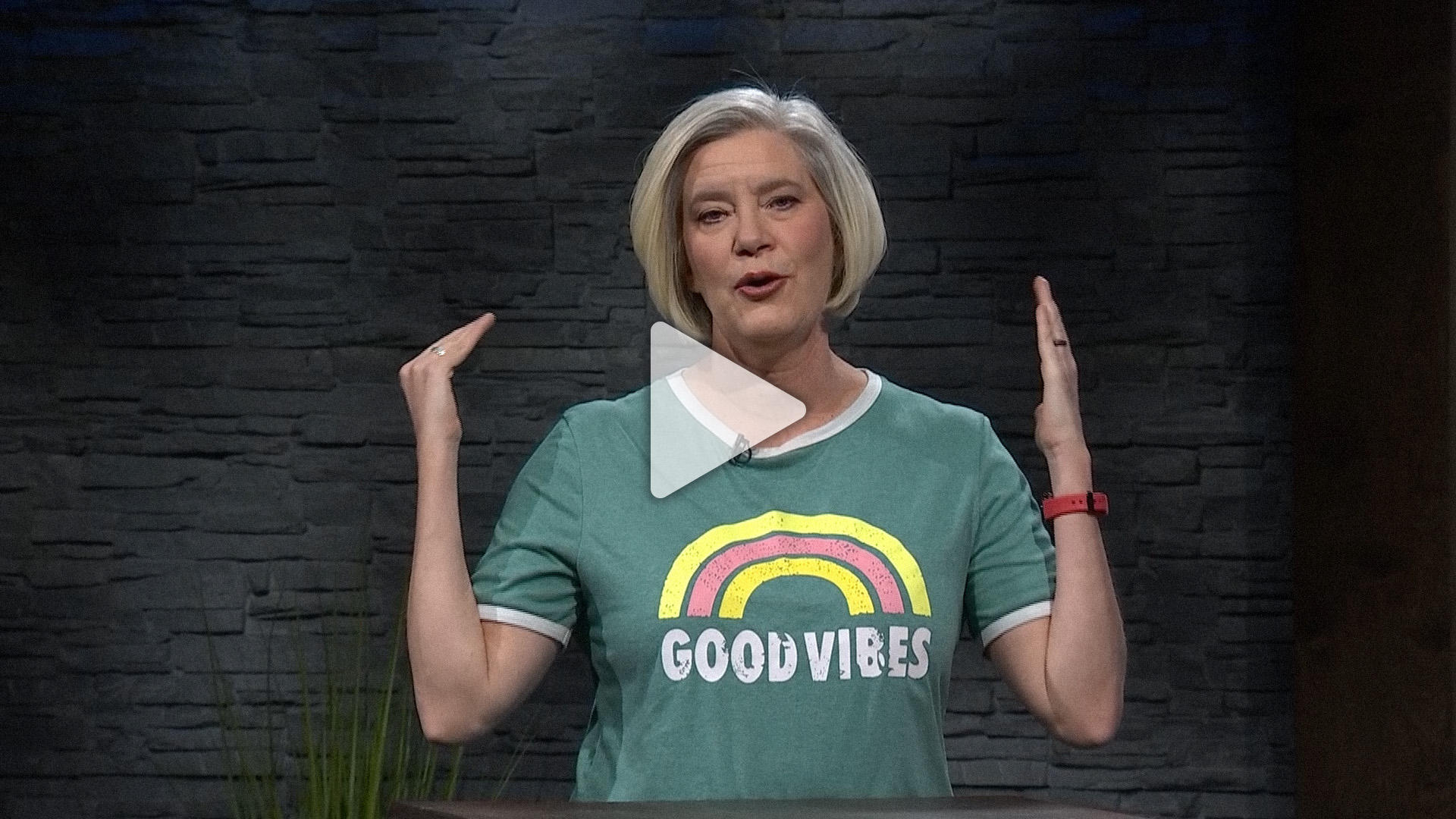 Woman wearing a "GOOD VIBES" t-shirt with a rainbow design, gesturing positively in a supportive environment, promoting gratitude and donations for vulnerable children.