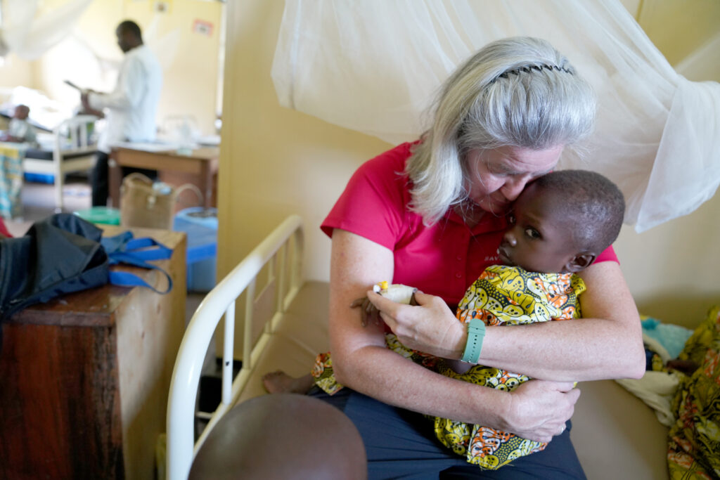 Woman in a red shirt holding malnourished child in a clinic, providing care and support, surrounded by medical staff and equipment, highlighting the mission of Saving Moses to combat malnutrition in Sub-Saharan Africa.