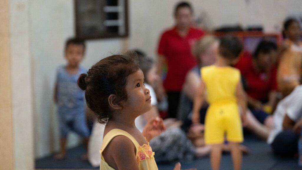 Young girl in a yellow outfit observing her surroundings, with children and caregivers in the background, illustrating the supportive environment of a NightCare Center for vulnerable infants and toddlers.