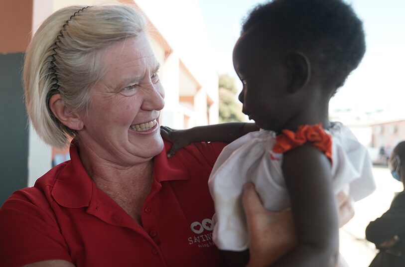 Woman smiling and holding an infant in a white dress, promoting maternal and infant health support through donations.