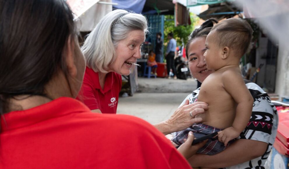 Woman in red shirt engaging with a baby held by a mother, smiling and interacting in a community setting, highlighting support for newborns and families.