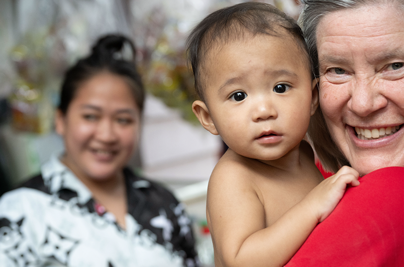 Smiling woman holding a baby, with a supportive background figure, highlighting care and connection in a context of helping vulnerable children.