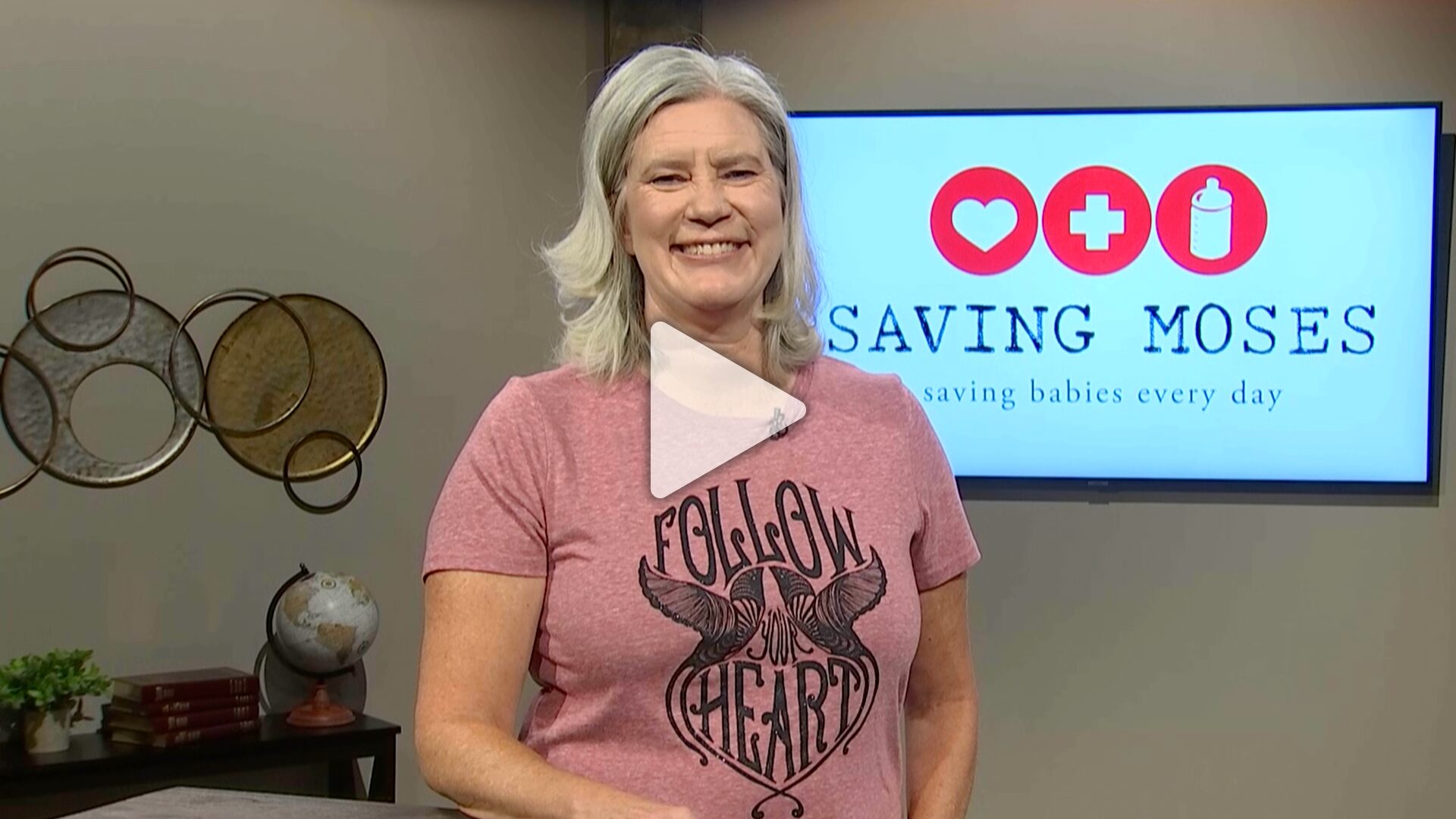 Smiling woman wearing a "Follow Your Heart" shirt, standing in front of a screen displaying the Saving Moses logo, emphasizing support for vulnerable babies and toddlers.