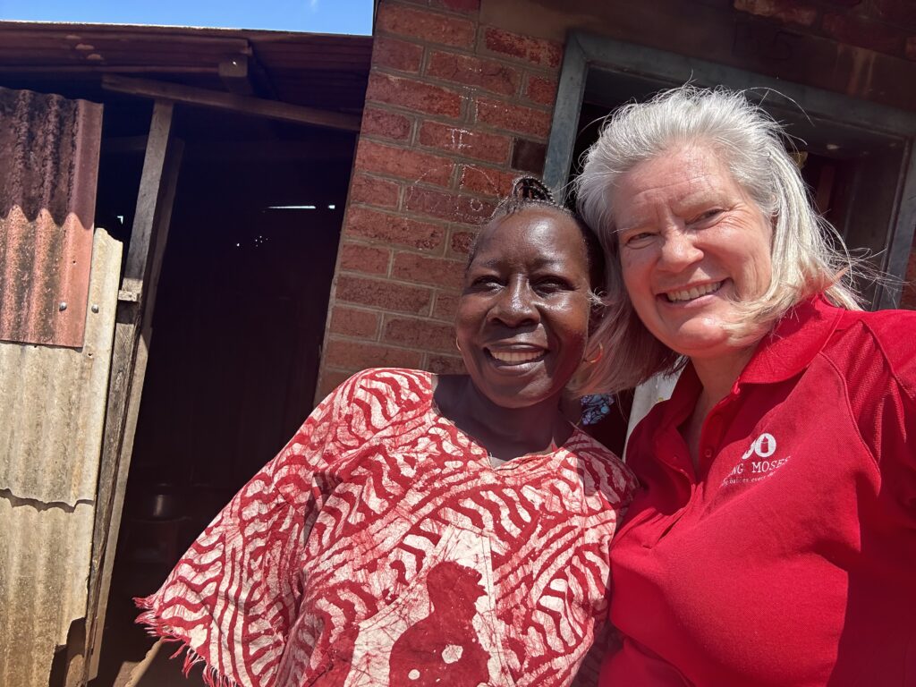 Two women smiling together outside a building, one wearing a red shirt with the Saving Moses logo and the other in a red patterned garment, representing community support and humanitarian efforts in regions served by Saving Moses.