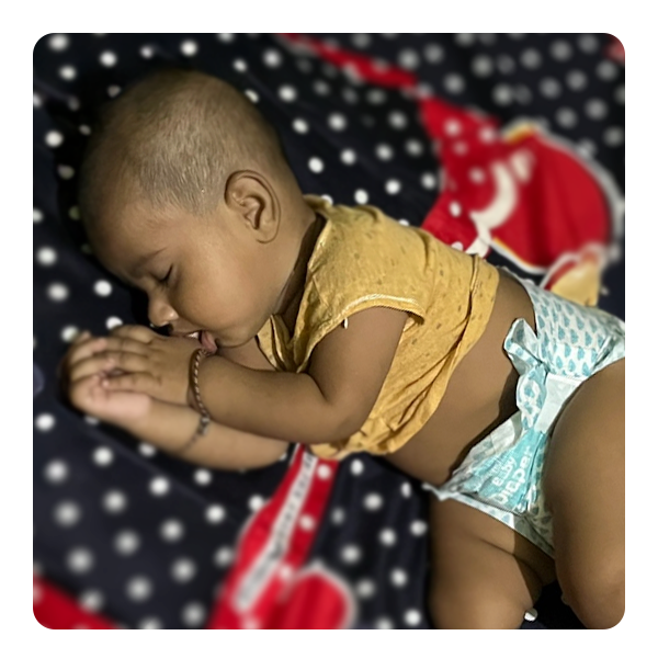 Infant peacefully sleeping on a bed, wearing a diaper and a yellow shirt, surrounded by a colorful patterned blanket, emphasizing the importance of safe sleep for vulnerable children.