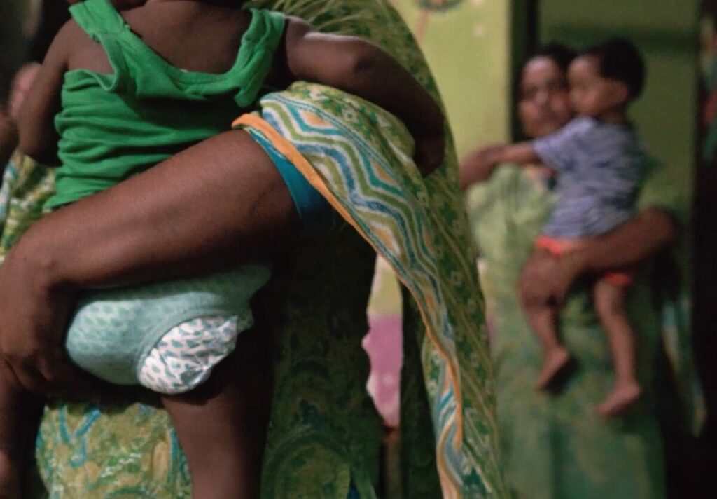 Mother holding a baby in a green outfit, surrounded by children in a dimly lit environment, illustrating the mission of Saving Moses to support vulnerable families.