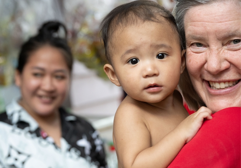 Smiling woman holding a baby in a supportive environment, with another woman in the background, highlighting the mission of Saving Moses to ensure safety for children of mothers in the sex industry.