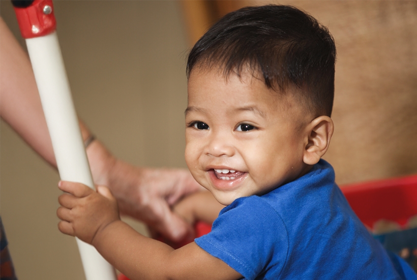 Smiling toddler in blue shirt holding onto a toy cart, representing hope and care for vulnerable children supported by Saving Moses.