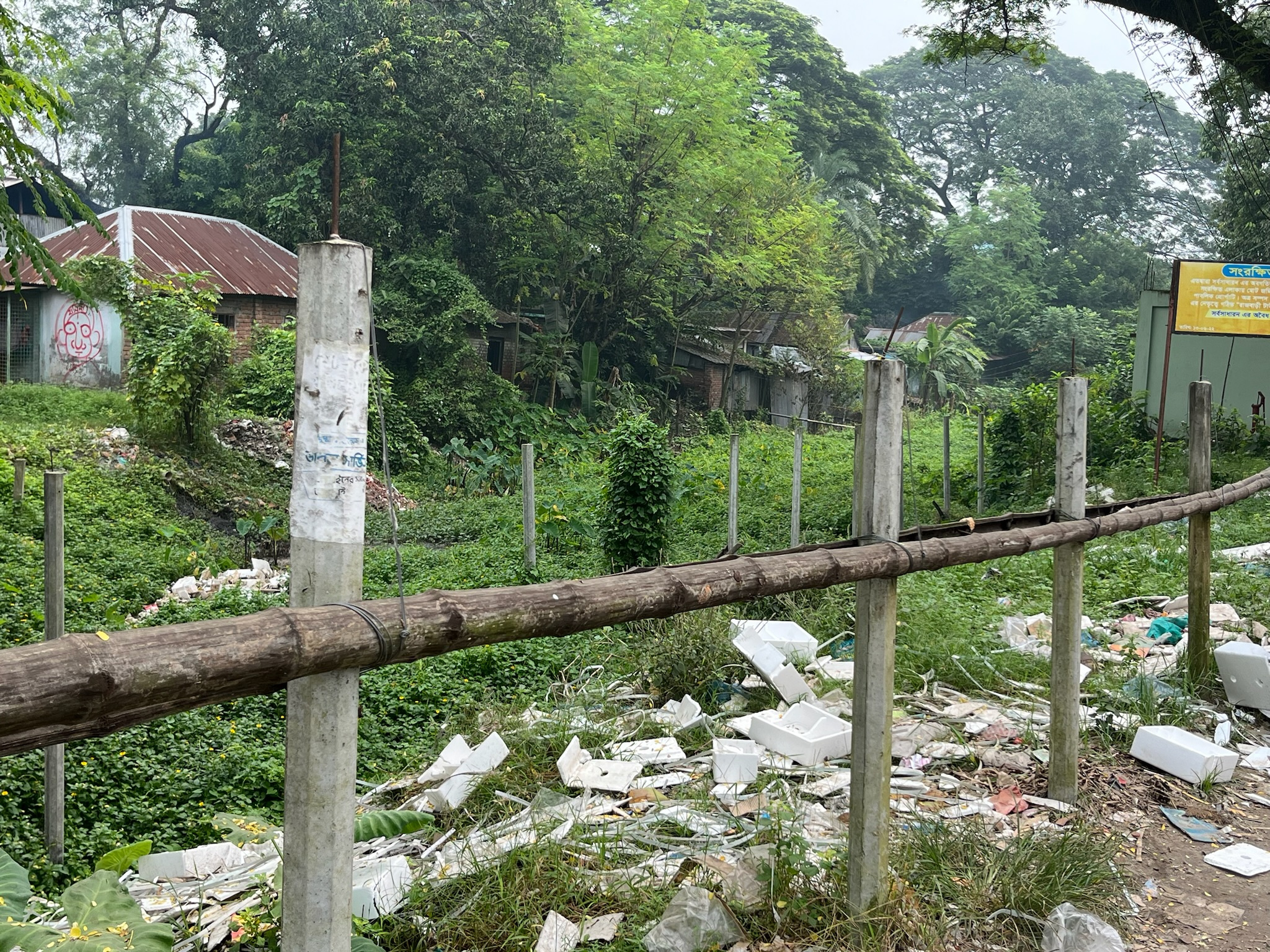 Littered landscape in Bangladesh with overgrown vegetation, wooden fence posts, and a backdrop of dilapidated structures, highlighting the challenging environment for vulnerable children supported by Saving Moses.