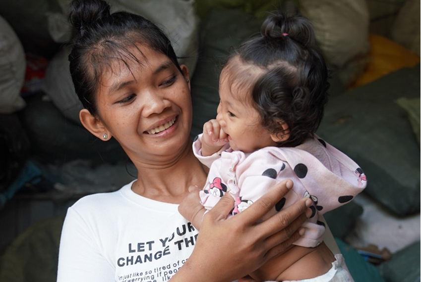 Smiling woman holding a baby in a pink polka dot hoodie, highlighting the bond of maternal care and support for newborns, relevant to Saving Moses' mission of ensuring safe childbirth and nurturing fragile lives.