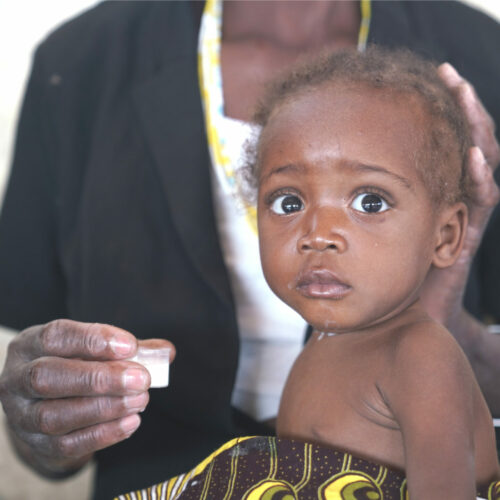 Child with large, expressive eyes, sitting on a caregiver's lap, holding a small container, representing the vulnerable infants supported by Saving Moses.