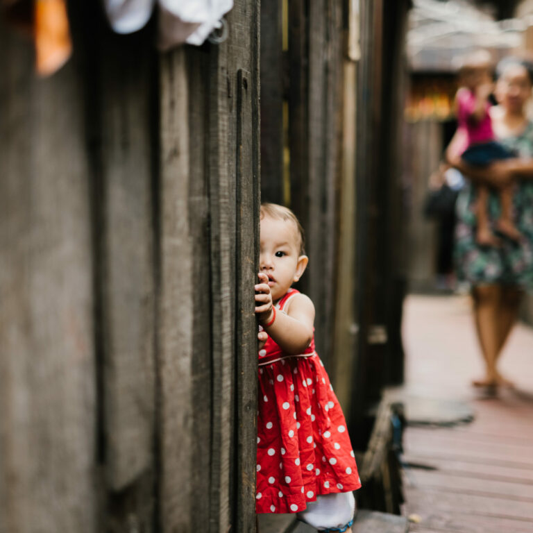 cambodia2016-169 Child in red polka dot dress peeking from between wooden planks, symbolizing vulnerability and innocence, relevant to Saving Moses' mission to protect babies and toddlers.